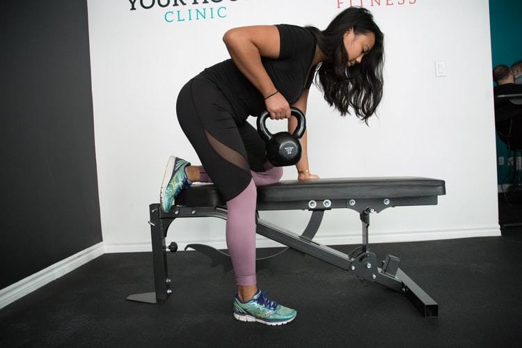 A woman lifting one kettlebell while her leg is resting on a flat table