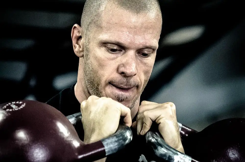 Man concentrating hard while each of his hands are lifting a pair of kettlebells