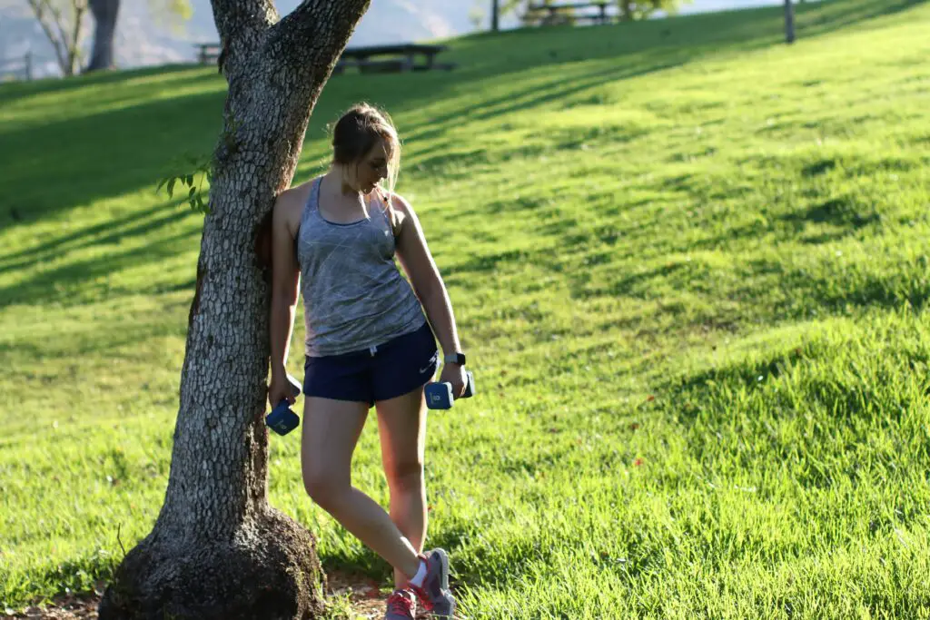 Woman holding a dumbbell with each hand, while leaning on a tree